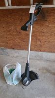 Full view of the Black and Decker string trimmer standing upright on concrete floor with a plastic bag containing trimmer line nearby.