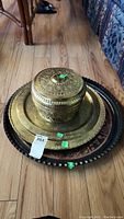 Photo showing stack of brass plates topped by a brass canister with lid, all with ornate engraved designs on the floor.