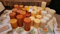 Wide view of the full assortment of pillar candles in various colors and sizes arranged on a cloth with a brick background.