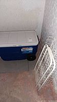 Blue Coleman cooler with white lid and black wheels, placed on floor corner.