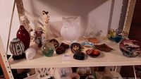 Overview of the shelf showing a collection of ceramic and glass figurines, bowls, vase, and decorative pieces including a white owl figurine and paperweights