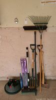 Photo showing metal leaf rake, wooden pitchfork, shovel, ax, push broom, dust pan, and wiper blades in packaging against a wall.