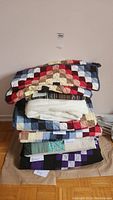 Stack of assorted bed textiles including multicolor patchwork quilts, grey pillow sham, white and striped bed wraps, floral quilt and black and purple quilt