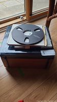 Wooden carrying case with large black metal tape spool on top by a window