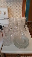 Clear glassware including a pitcher, multiple footed coffee mugs with handles, and shrimp cocktail glasses arranged on a countertop.