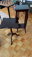 Two dark wood side tables positioned near each other on a parquet wood floor. The pedestal table with tripod legs in foreground and the taller table with lower shelf in background.