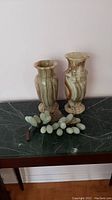 Two onyx vases and stone grapevine decoration displayed on dark green marble surface.