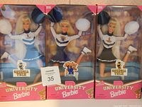 Photo of three University Barbie dolls in original packaging. One Kentucky Barbie and two Georgia Tech Barbies, each in cheerleading uniforms with pom-poms.