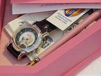 Close-up photo showing Barbie wristwatch with black leather strap, watch face has Barbie image and logo, accompanied by a small folded leaflet and pink pen; items are new in packaging.