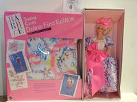 Classic Barbie doll in box wearing floral puffed sleeve dress with large pink bow, alongside boxed Barbie Deluxe First Edition Trading Cards