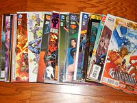 Photo of over 25 comic books placed side by side on a wooden floor, showing covers from multiple publishers including DC, Marvel, IDW, and Dynamite. Comics are in protective sleeves and in mixed condition.