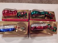 Four glass shoe-shaped ornaments in red, green, blue, and gold colors, each paired with a miniature Hot Wheels car inside clear plastic cases on gold backing within original boxes. All items are new in box.