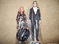 Two dolls, one male in tuxedo and one female in colorful black dress, both wrapped in plastic, laid on carpeted floor.