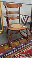Front angled view of antique wooden rocking chair showing embossed leather seat bordered with nail heads, carved wooden backrest arms and spindles pattern.