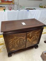 View of dark wood Asian cabinet front showing two doors with detailed gold-tone landscape designs, metal ring handles, flat top and sculpted legs.