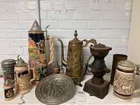 Wide view showing the full set of seven uniquely decorated beer steins, brass coffee grinder, brass vase, small iron cast item, and decorative metal plate arranged on surface against brick wall.