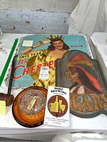 Photo showing vintage cardboard Chesterfield advertisement sign with a woman holding cards, a carved wooden Chief sign, glass ashtray with amber base, Home Brewing book, and a pipe with note