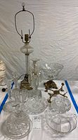Wide shot showing 29.5 inch glass lamp with brass fittings and multiple pieces of clear cut glass including bowls, dishes, and candle holders on a white cloth.