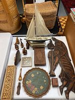 Overview of all items on table: model sailboat, angel wood wall carving, multiple wood animal carvings, decorative wooden boxes, floral plate, small wooden utensils.