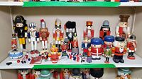 Wide view of multiple wooden Nutcracker and toy soldier figurines, varying sizes, some showing damage and missing parts, arranged on white shelves.