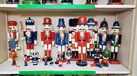 Seventeen wooden nutcracker figurines displayed on shelving with varying sizes and colors, showing some with loose or missing parts.