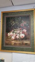 Full view of the framed floral oil painting showing basket of pink roses and dark foliage background, gold decorative frame, mounted on beige wall.