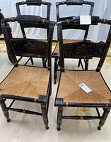 Front view of vintage Hitchcock chairs showing black lacquered wood with gold stenciled decorative accents and rush woven seats. One chair has visible signature.