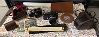 Wide shot showing both vintage cameras, multiple cases including a round brown filter case, booklets/manuals, and other camera accessories on a floral tablecloth.