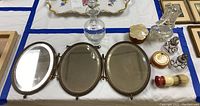 Top view of the fully opened 3-tier folding mirror, large perfume decanter, smaller perfume bottle, jars, and brushes laid out on a white cloth.