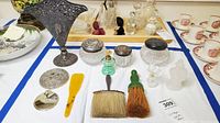 Overview of vintage vanity items laid out on table including grooming brushes, glass jars with metal lids, perfume bottles, shoe horn and filigree vase