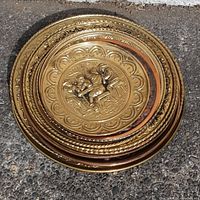 Stacked view of multiple decorative brass and copper plates showing different sizes and embossed designs with a central embossed figure and decorative border patterns