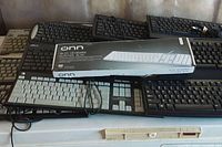 12 computer keyboards stacked, including an ONN slim keyboard in original packaging on top.