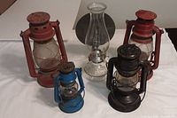 Five hurricane barn lamps in various colors and sizes, showing rust spots, peeling paint, and one clear glass lamp with metal base.