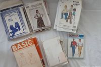 Multiple stacks of assorted sewing patterns from brands Denim, Simplicity, Kwik Sew, Burda, and Basic Knits, both cut and uncut patterns visible