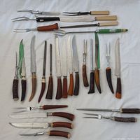 Top-down view of 26 carving knives and forks with various bone and Bakelite handles laid on white cloth background.