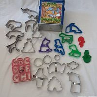 Various cookie cutters including animal shapes in metal and colorful plastic alphabet cutters in a grid on white fabric.