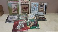 Photo of 10 cross stitch and stitchery kits arranged on a floor, showing packaging with various designs including birds, angels, stockings, Winnie the Pooh, butterflies, and scenic scenes.