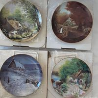 Four Dawn Karr Olde Country Cottages collector plates, viewed from above in their packaging with box lids underneath. Each plate depicts a different rustic cottage scene.