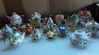 14 whimsical novelty teapots displayed together on a light surface, featuring different colors and shapes including cottage-shaped teapots and floral designs.