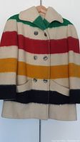 Front of vintage Hudson Bay wool jacket with green, red, yellow, black stripes and silver buttons, hanging on hanger