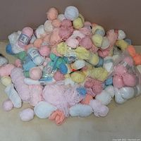 Photo showing a large pile of assorted baby yarn balls in various pastel colors piled on a beige surface. Some yarn balls still have labels, others are loose and partially unwound.