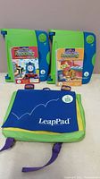 Two Leap Pad Learning systems with two books and cartridges and a LeapPad branded backpack carrying case on a light background.