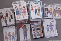 Photo of multiple stacks of sewing pattern packets spread on floor, packaging showing various children’s and youth clothing designs from brands including Butterick, McCall's, Simplicity, Burda.