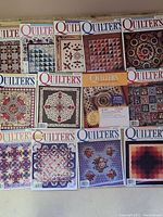 Photo showing 12 Quilter's Newsletter Magazine issues with quilt pattern covers featuring geometric, floral, and traditional quilt block designs in various colors.