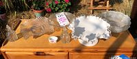 Lot of glass platters, bowls, bottles, and a small egg cup arranged on a wooden surface outside in sunlight.