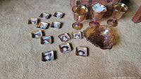 Full set of carnival glass goblets, bowl, and twelve small decorative dishes with painted Victorian women's faces arranged on carpeted floor.