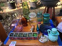 Overview of all colored glass items on a wooden table outdoors, showing green vases, jars, blue bowl, corked pitcher, ice cube trays, and other glass pieces