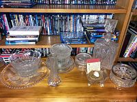 Lead crystal decanter with stopper, several plates, dishes, and a small vase arranged on wooden surface with books behind.