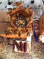 Front view of the cuckoo clock showing carved leaf decoration, clock face with Roman numerals, and three pine cone-shaped weights on the ground in front.