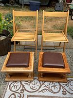 Front view of two wooden folding chairs and two wooden footstools with brown leather cushions, arranged outdoors on a patterned rug. Visible signs of wear on footstools.